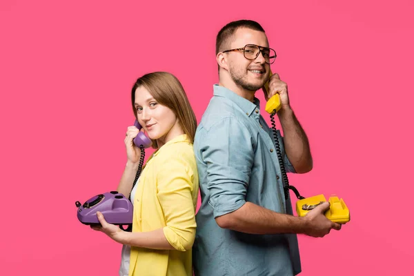 Seitenansicht eines glücklichen jungen Paares, das Rücken an Rücken steht und mit Drehtelefonen spricht, isoliert auf rosa — Stockfoto
