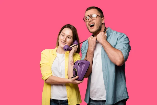Lächelnde junge Frau, die per Hörer spricht, während Mann mit Draht um den Hals isoliert auf rosa schreit — Stockfoto