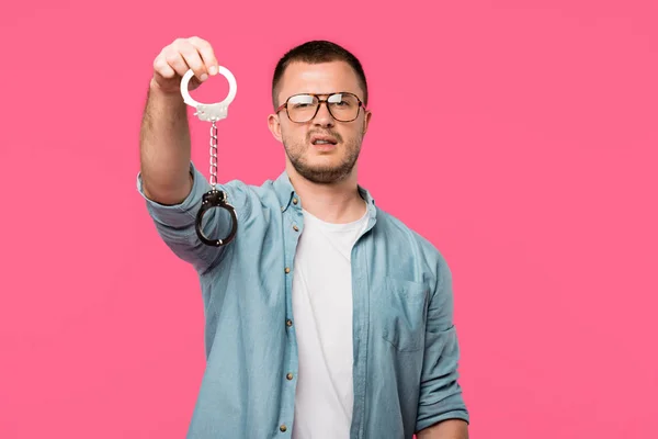Молодий чоловік в окулярах тримає наручники і дивиться на камеру ізольовано на рожевому — стокове фото