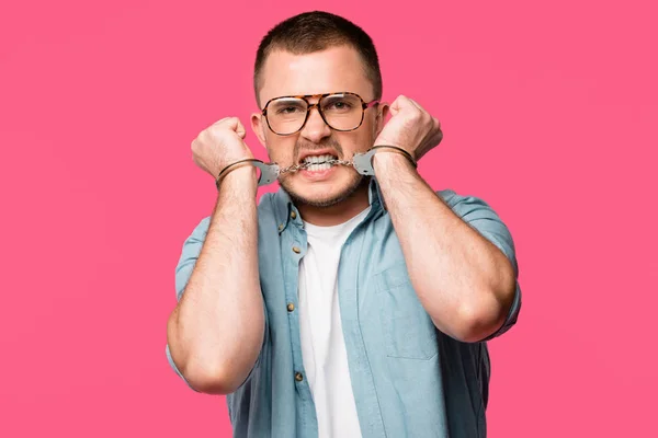 Aggressive young man in eyeglasses biting handcuffs and looking at camera isolated on pink — Stock Photo