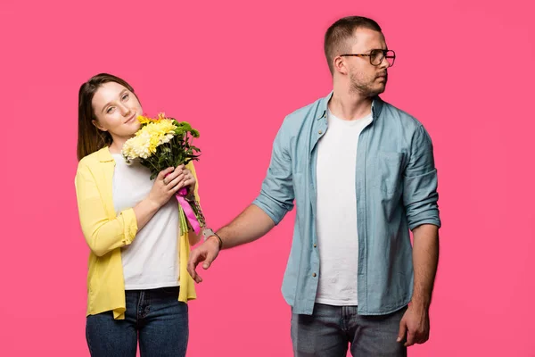 Smiling young woman holding bouquet of flowers while standing in handcuffs with man looking away isolated on pink — Stock Photo