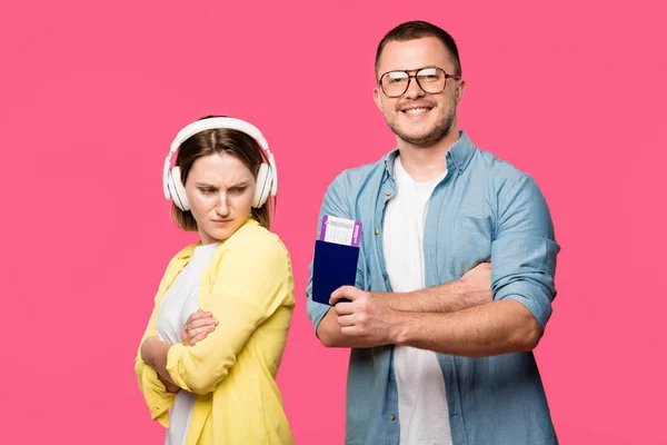 Upset woman in headphones looking at happy man in eyeglasses holding passport and boarding pass isolated on pink — Stock Photo
