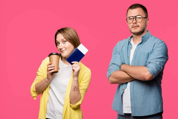 Mann mit Brille steht mit verschränkten Armen und blickt in die Kamera, während lächelnde Frau Pass, Bordkarte und Coffee to go auf rosa — Stockfoto