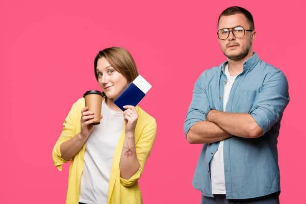 Uomo in occhiali in piedi con le braccia incrociate e guardando la fotocamera mentre sorridente donna in possesso di passaporto, carta d'imbarco e caffè per andare isolato su rosa — Foto stock