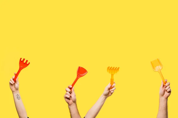 Cropped shot of male and female hands holding red and yellow plastic shovels and rakes isolated on yellow — Stock Photo