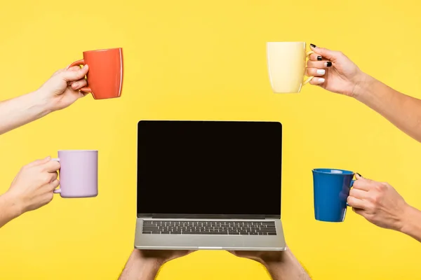 Recortado tiro de manos sosteniendo tazas y portátil con pantalla en blanco aislado en amarillo - foto de stock