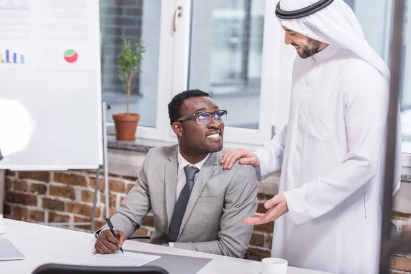 Afrikanisch-amerikanischer Geschäftsmann hält Stift in der Hand und blickt auf arabischen Partner im Amt — Stockfoto