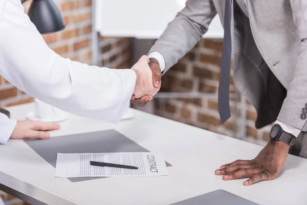 Мультиэтнические бизнесмены пожимают руки перед подписанием контракта в офисе — стоковое фото