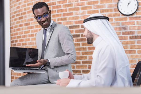 Un homme d'affaires afro-américain montre un ordinateur portable à un partenaire arabe au bureau — Photo de stock