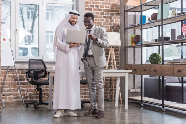 Des hommes d'affaires multiethniques debout avec ordinateur portable dans un bureau moderne — Photo de stock