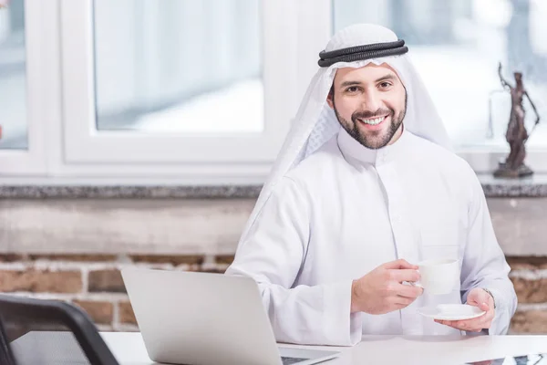 Homem de negócios árabe bebendo café perto de laptop no escritório — Fotografia de Stock