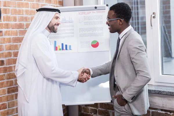 Empresários multiétnicos apertando as mãos perto de placa branca no escritório — Fotografia de Stock