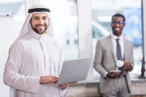 Arabian businessman holding laptop in office with african american partner on background — Stock Photo