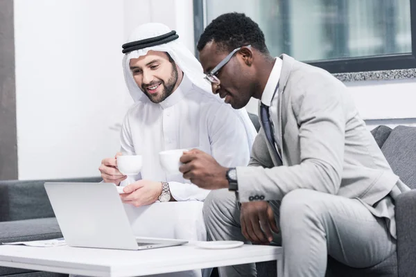 Empresarios multiculturales sosteniendo copas y mirando el portátil en la oficina moderna - foto de stock