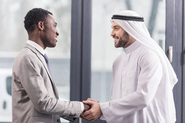 Close up view of african american businessman and arabic partner smiling and shaking hands in office — Stock Photo