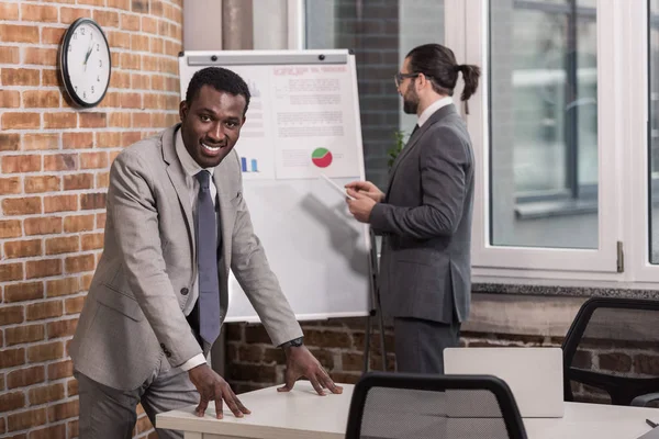 Hombre de negocios afroamericano mirando a la cámara y al socio de pie frente al rotafolio - foto de stock