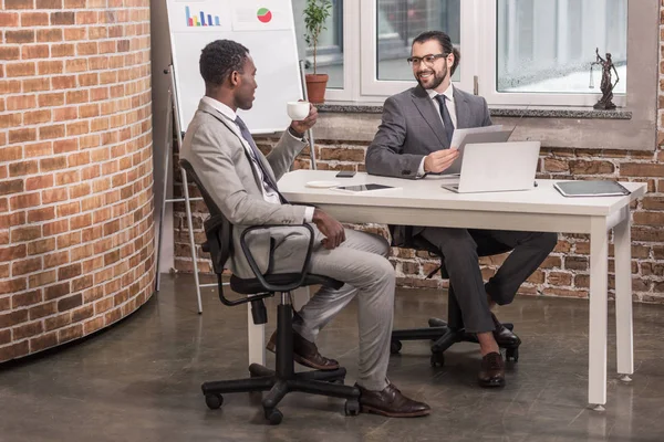 Multicultural business partners sitting at office table, drinking coffee and having discussion — Stock Photo