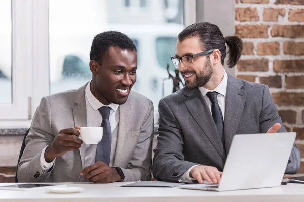 Улыбающиеся мультикультурные бизнесмены пьют кофе и смотрят на ноутбук в офисе — стоковое фото