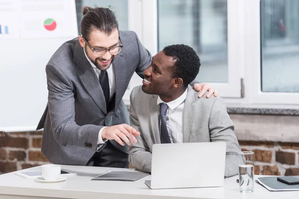 Красивий бізнесмен обіймає афроамериканського партнера і вказує на ноутбук в офісі — стокове фото