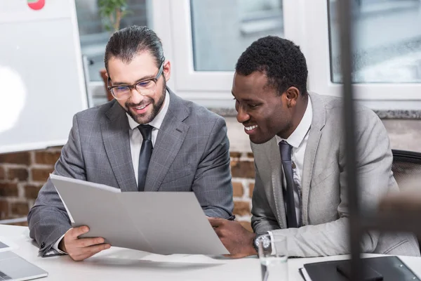 Мультикультурные деловые партнеры читают документы и улыбаются в офисе — стоковое фото