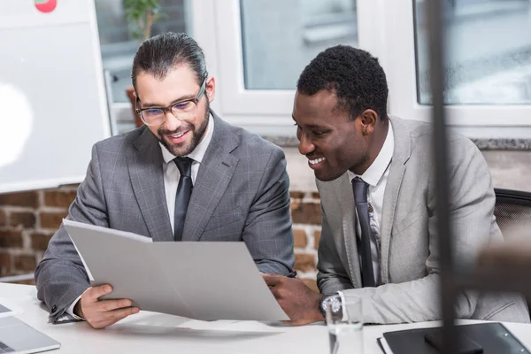 Socios comerciales multiculturales leyendo documento y sonriendo en la oficina - foto de stock