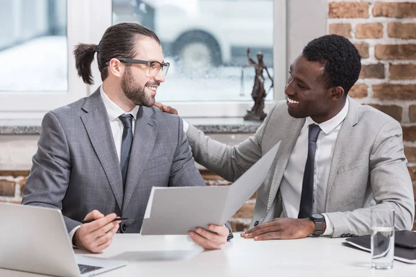 Smiling multicultural business partners sitting at table looking at each other in office — Stock Photo