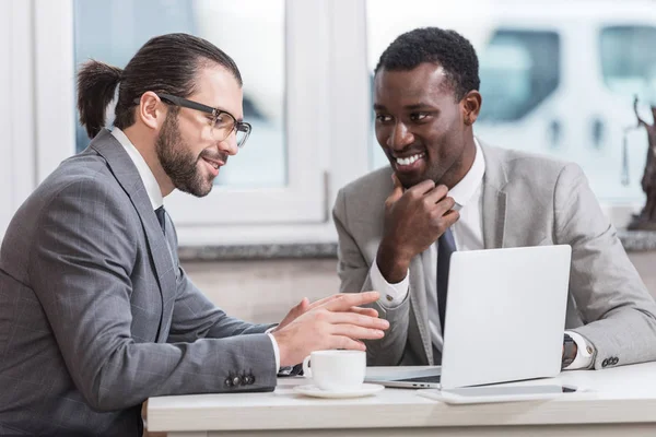 Uomini d'affari multiculturali sorridenti seduti a tavola con il computer portatile e avendo discussione — Foto stock