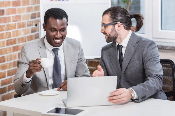 Bonito empresário apontando para laptop e sorrindo parceiro afro-americano beber café no escritório moderno — Fotografia de Stock