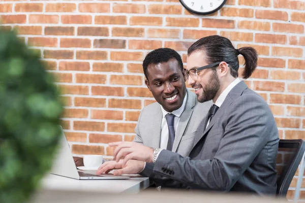 Gutaussehende multikulturelle Geschäftsleute lächeln und sitzen mit Laptop am Tisch — Stockfoto