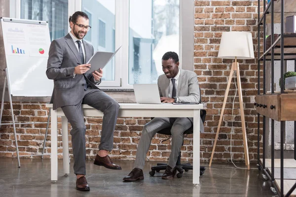 Усміхаючись бізнесмен, сидячи на таблиці та читання документа під час афро-американських партнера, що працюють на ноутбук в officeac — стокове фото