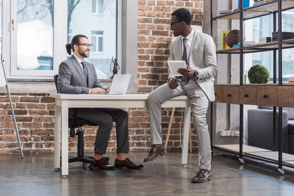 Gutaussehender Geschäftsmann sitzt am Tisch und schaut seinen afrikanisch-amerikanischen Partner mit digitalem Tablet im Büro an — Stockfoto