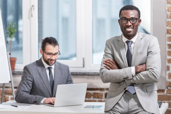 Afrikanisch-amerikanischer Geschäftsmann mit verschränkten Armen blickt in die Kamera, während sein Partner im Hintergrund am Laptop arbeitet — Stockfoto