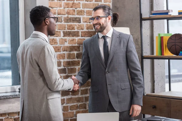 Улыбающийся красивый бизнесмен и африканский американский партнер пожимающий руки в офисе — стоковое фото