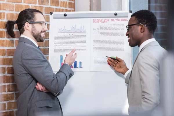 Smiling multicultural businessmen talking in front of white board with charts and graphs in office — Stock Photo