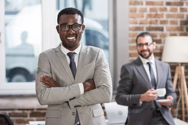 Sonriente hombre de negocios afroamericano con los brazos cruzados mirando a la cámara y apuesto compañero de fondo - foto de stock