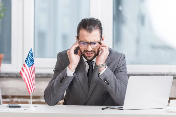 Hombre de negocios decepcionado sentado en el escritorio de la oficina con el ordenador portátil y la bandera americana - foto de stock