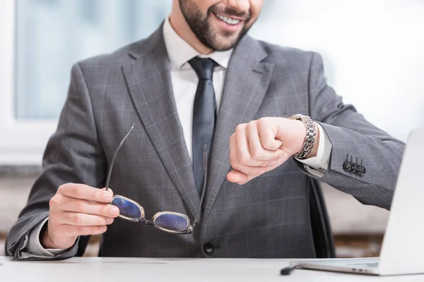 Cropped view of smiling businessman holding glasses and looking at watch — Stock Photo