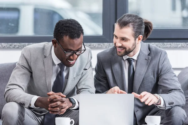 Lächelnde multikulturelle Geschäftsleute in Anzügen sitzen mit Laptop am Tisch im Büro — Stockfoto
