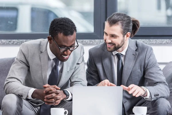 Hommes d'affaires souriants multiculturels en costumes assis à la table avec ordinateur portable dans le bureau — Photo de stock