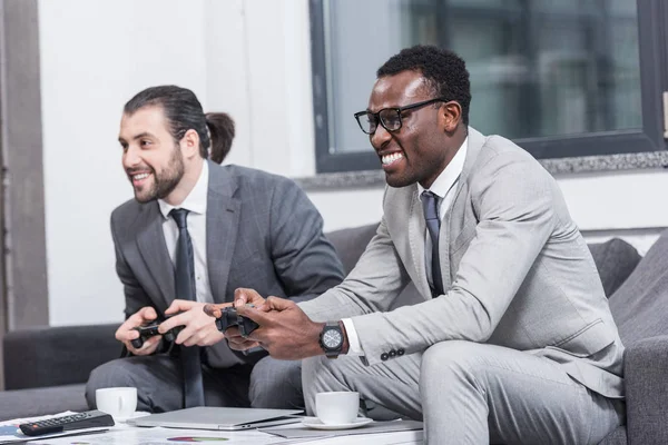 Multiethnic businessmen sitting on couch and playing video game with gamepads — Stock Photo