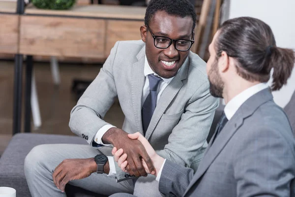 Multiethnic business partners sitting on sofa and shaking hands — Stock Photo