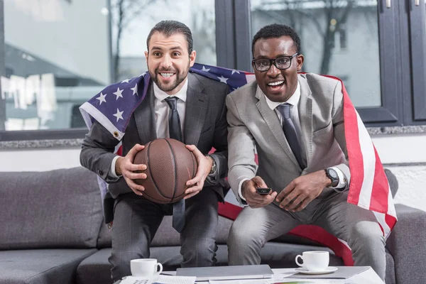 Multiethnic businessmen with american flag on shoulders watching basketball match in office — Stock Photo