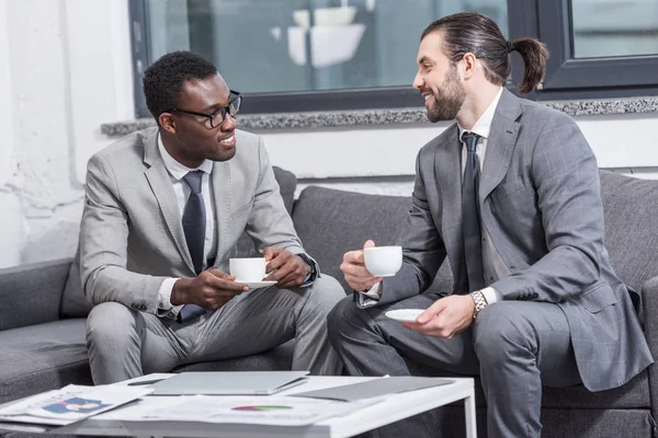 Smiling multicultural businessmen sitting on couch, looking at each other and drinking coffee in office — Stock Photo