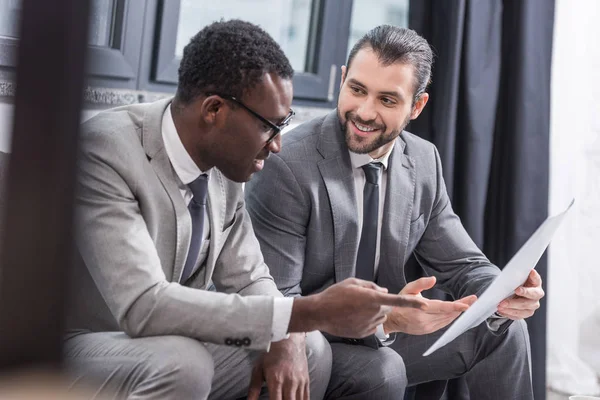 Empresarios multiétnicos sentados en el sofá y discutiendo el documento de negocios - foto de stock