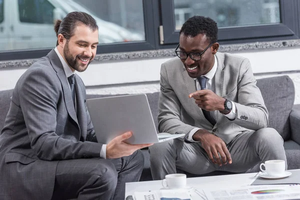 Hombre de negocios afroamericano sentado en el sofá y señalando con el dedo a los socios de negocios portátil en la oficina - foto de stock