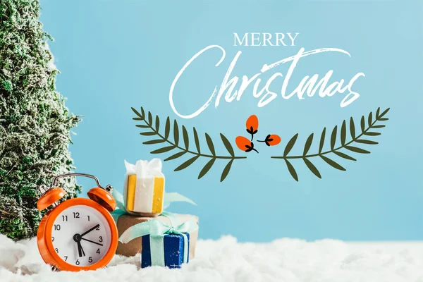 Primer plano de regalos de Navidad con despertador y árbol de Navidad en miniatura de pie sobre nieve sobre fondo azul con letras de 