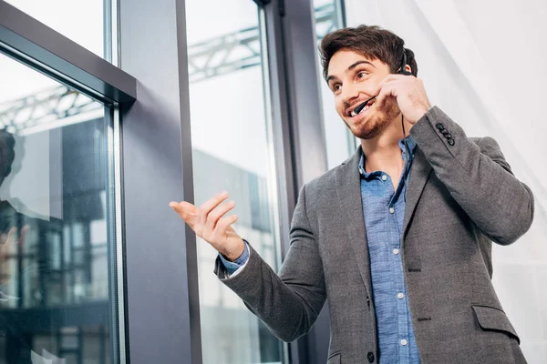 Schöner Callcenter-Betreiber, der am Fenster steht, Headset anfasst und im Büro in Mikrofon spricht — Stockfoto