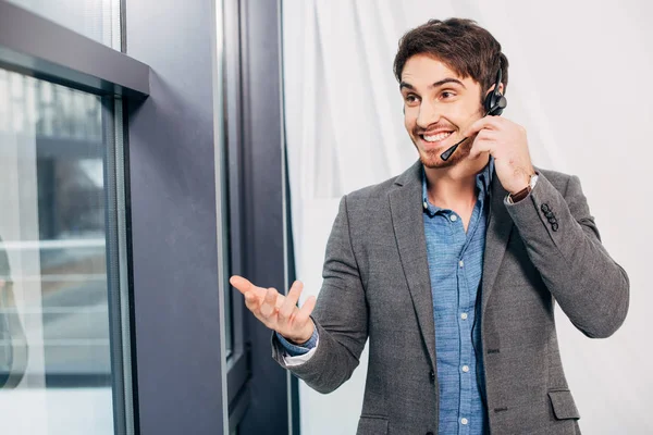 Sonriente operador del centro de llamadas de pie junto a la ventana y hablando en micrófono en la oficina - foto de stock
