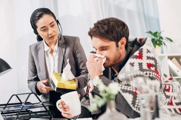 Inquieto operador del centro de llamadas dando servilletas a compañero de trabajo enfermo con manta y taza en la oficina - foto de stock