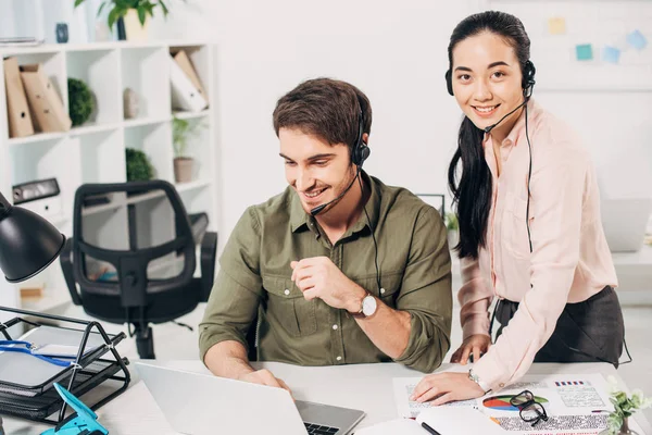 Sorrindo operador de call center trabalhando no laptop e muito colega de trabalho olhando para a câmera no escritório — Fotografia de Stock